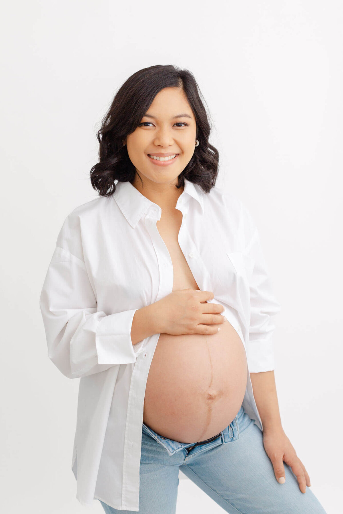 Mama-to-be with shoulder length black hair in loose curls. She is wearing a white buttoned down shirt that is open and showing off her baby bump. She has light colored jeans on that have the buttons open at the top.