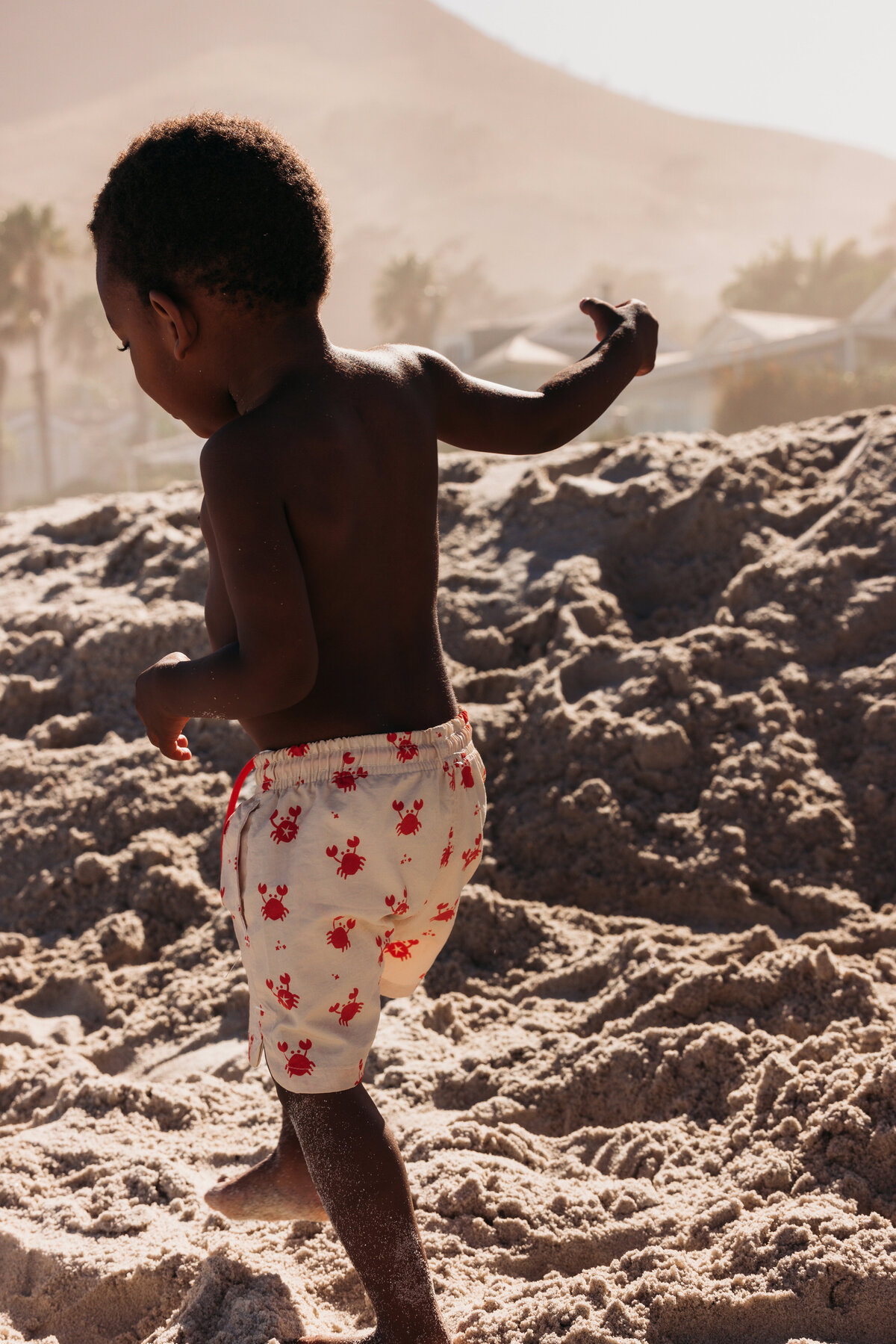 Young boy walking through soft beach sand in printed crab swim shorts, captured mid-motion with warm sunlight and a hazy mountain in the background.