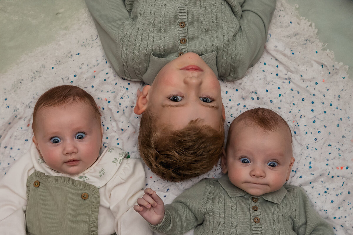 Three children lying down, with a toddler in the center flanked by twin babies dressed in green, all staring upward at the camera.