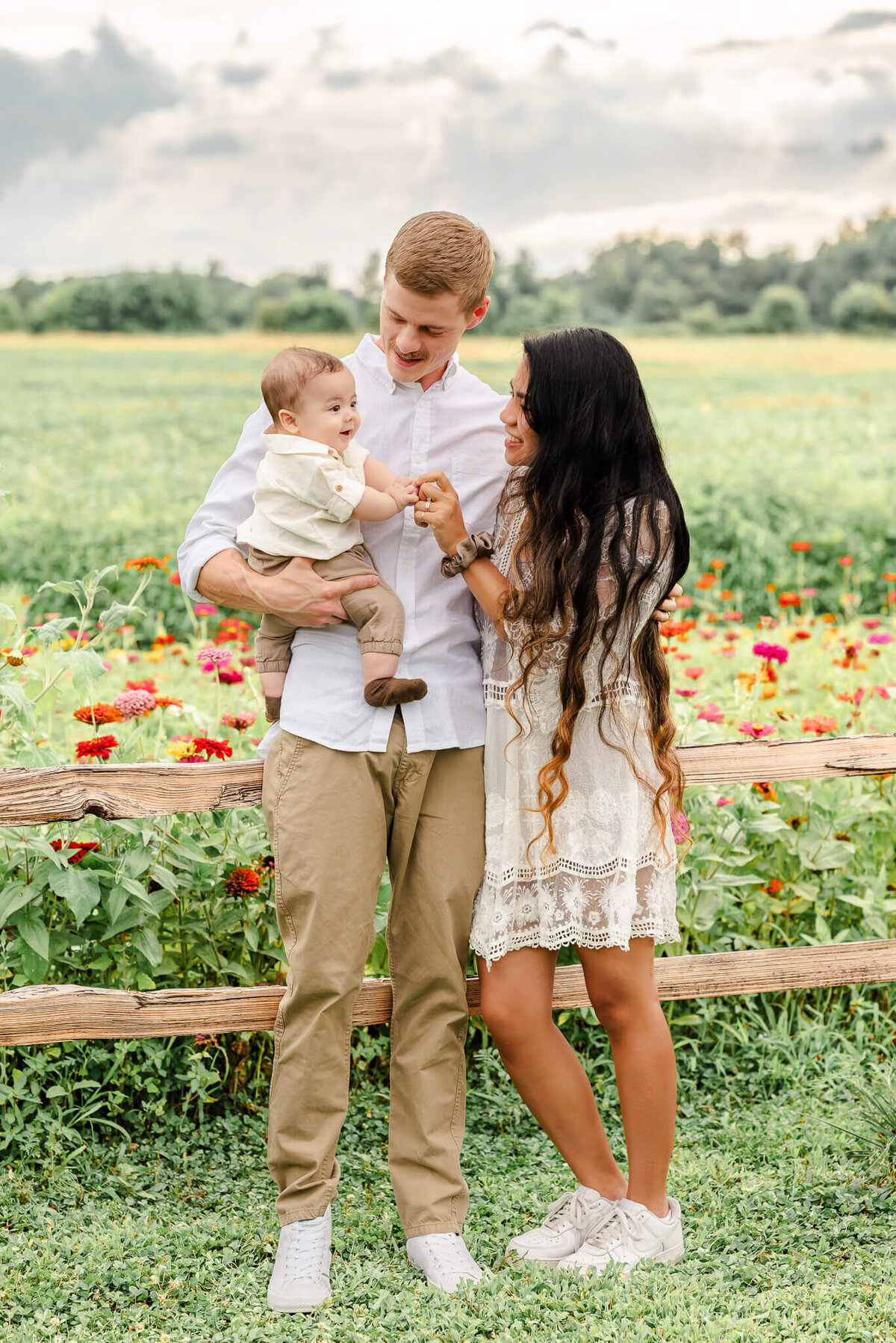 A young couple, wearing neutrals, look at their infant son lovingly while having family pictures done in a flower field.
