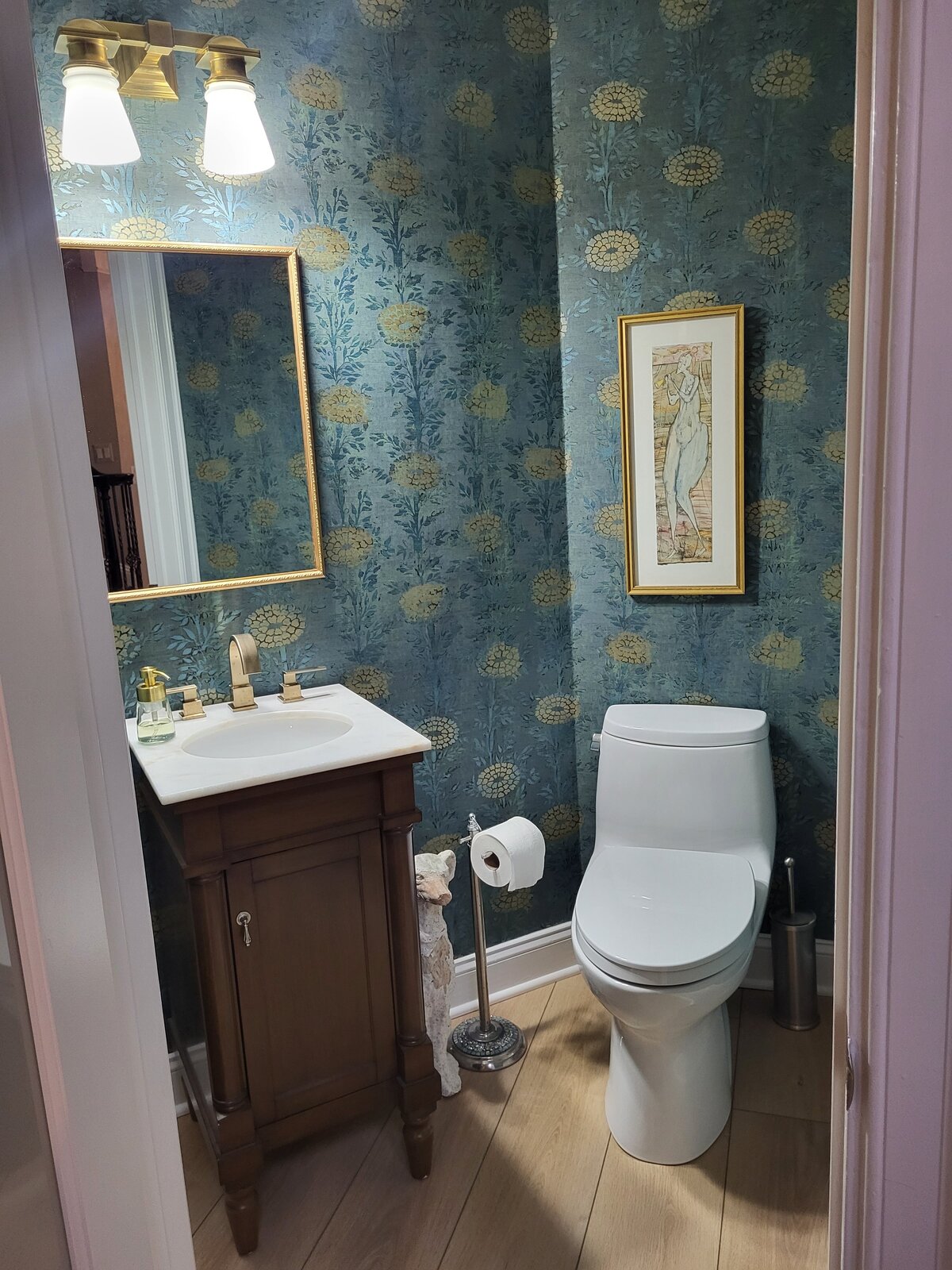 Wallpapered bathroom with teal background with golden chrysanthemum  with small vanity with a brass mirror and lighting