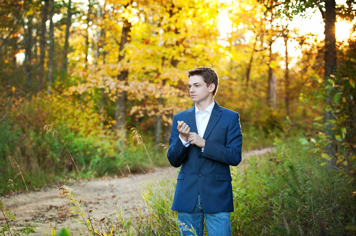 High School senior guy standing next to gravel road with fall colors behind him rolling up sleeve to blue blazer jacket.