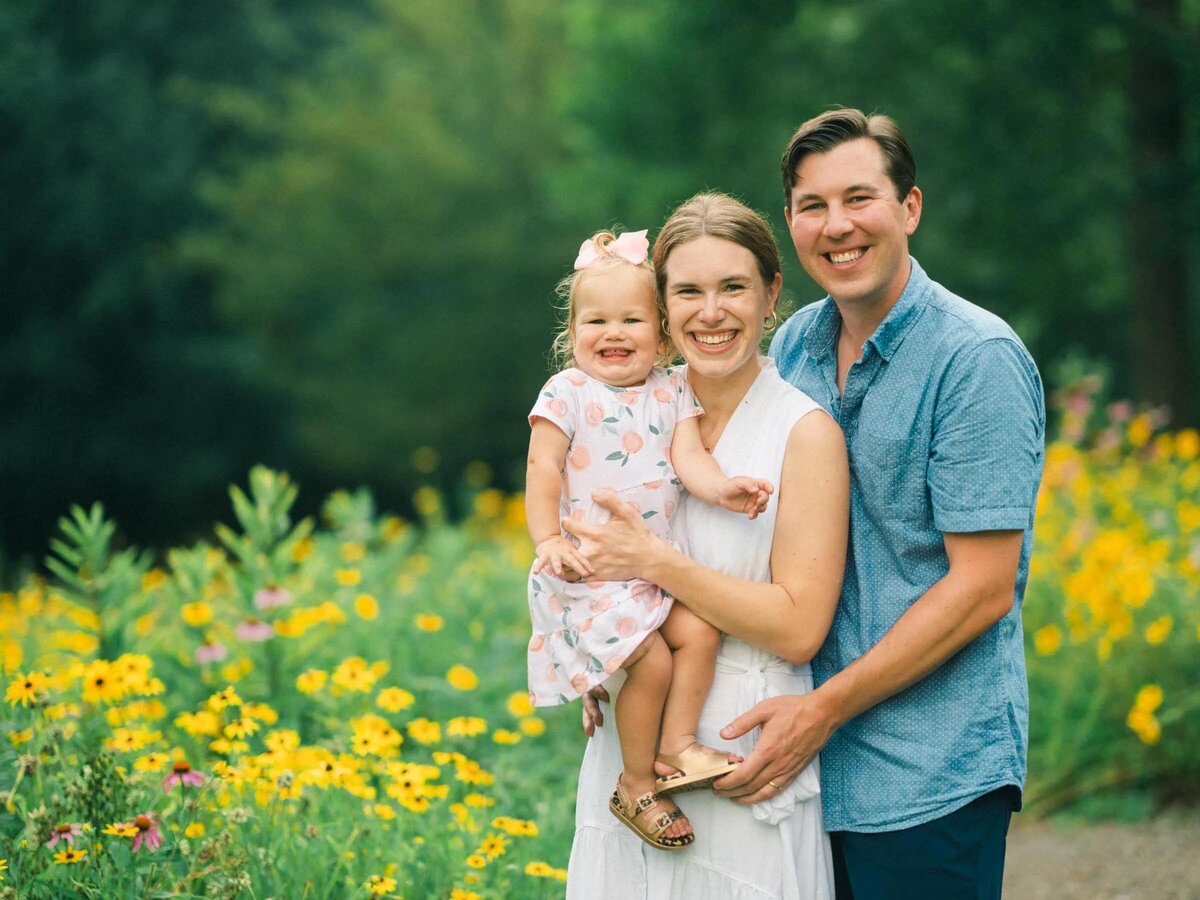 A man standing behind his partner as she holds their child.