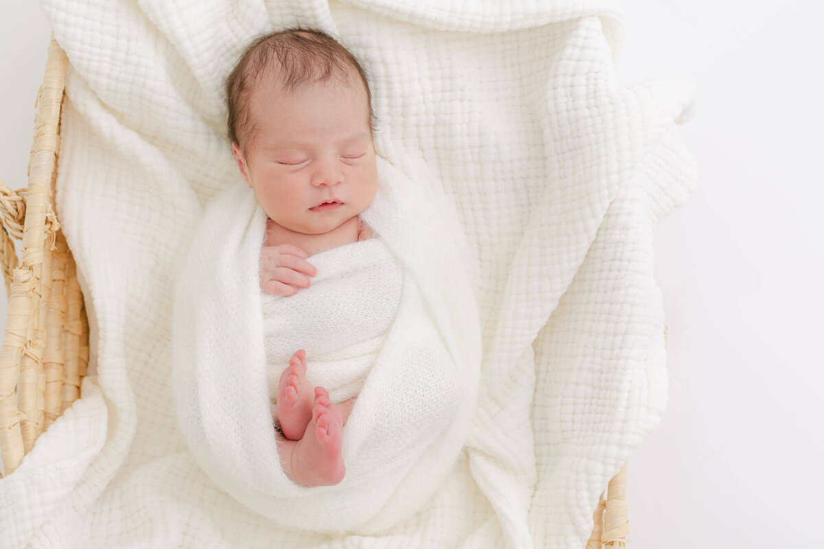 baby swaddled in white and sleeping in a moses basket on top of some white blankets.