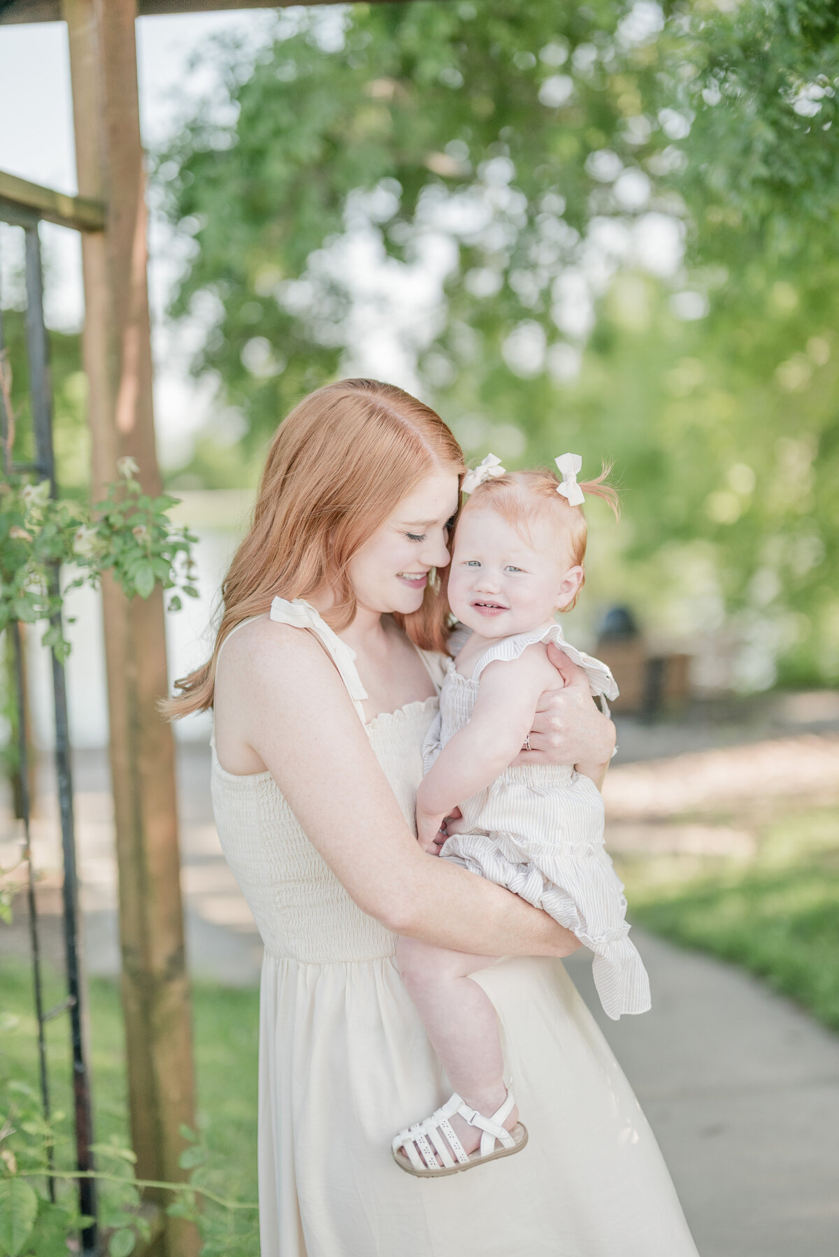 Mother holding and nuzzling her toddler daughter near a rustic fence in an outdoor park setting.