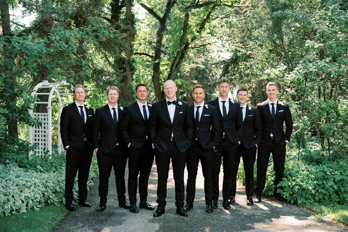 A group of eight men in black suits and ties stand on a garden path at the Norland Historic Estate, posing for a wedding photograph. Trees and greenery surround them, capturing the elegance of a wedding in Calgary.