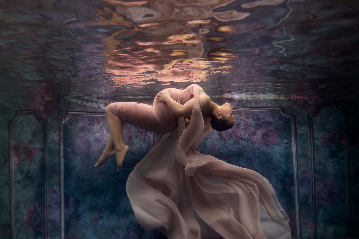 A breathtaking underwater maternity photo by a Dallas underwater pregnancy photographer, capturing the elegance of a woman floating gracefully in a soft pink gown. Her body arches backward, supported by the water, while sheer fabric flows around her like delicate petals. The dreamy floral backdrop adds to the serene, artistic composition of this underwater maternity photoshoot.
