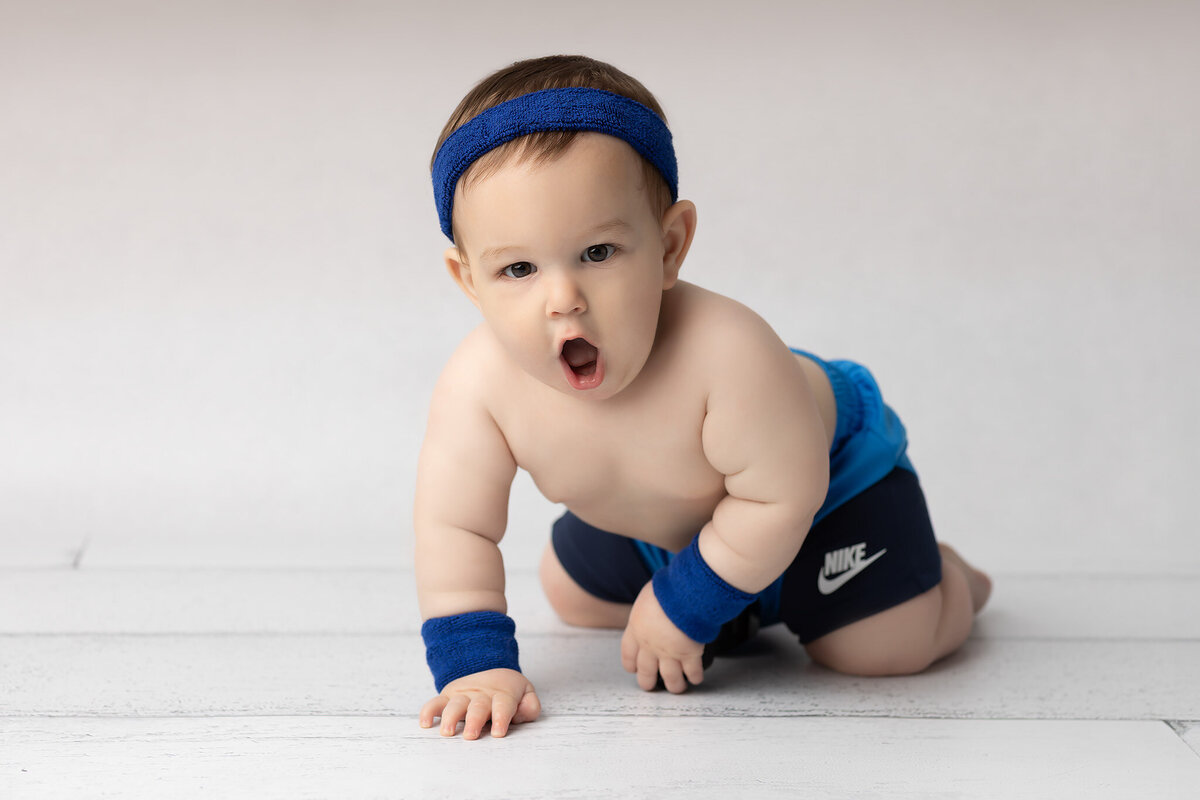 Crawling baby boy in blue Nike shorts, sweatbands, and a headband strikes a playful pose with an open-mouthed expression during a Dallas milestone photography session on a white floor.