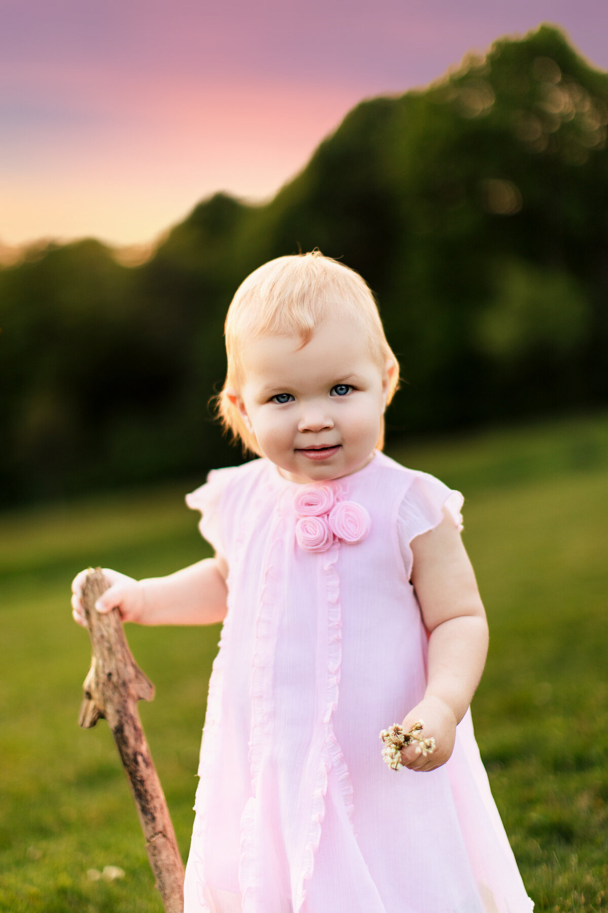 Three year old infant girl holds a walking stick and flowers in the woods during a family portrait session near Sewickley Pennsylvania