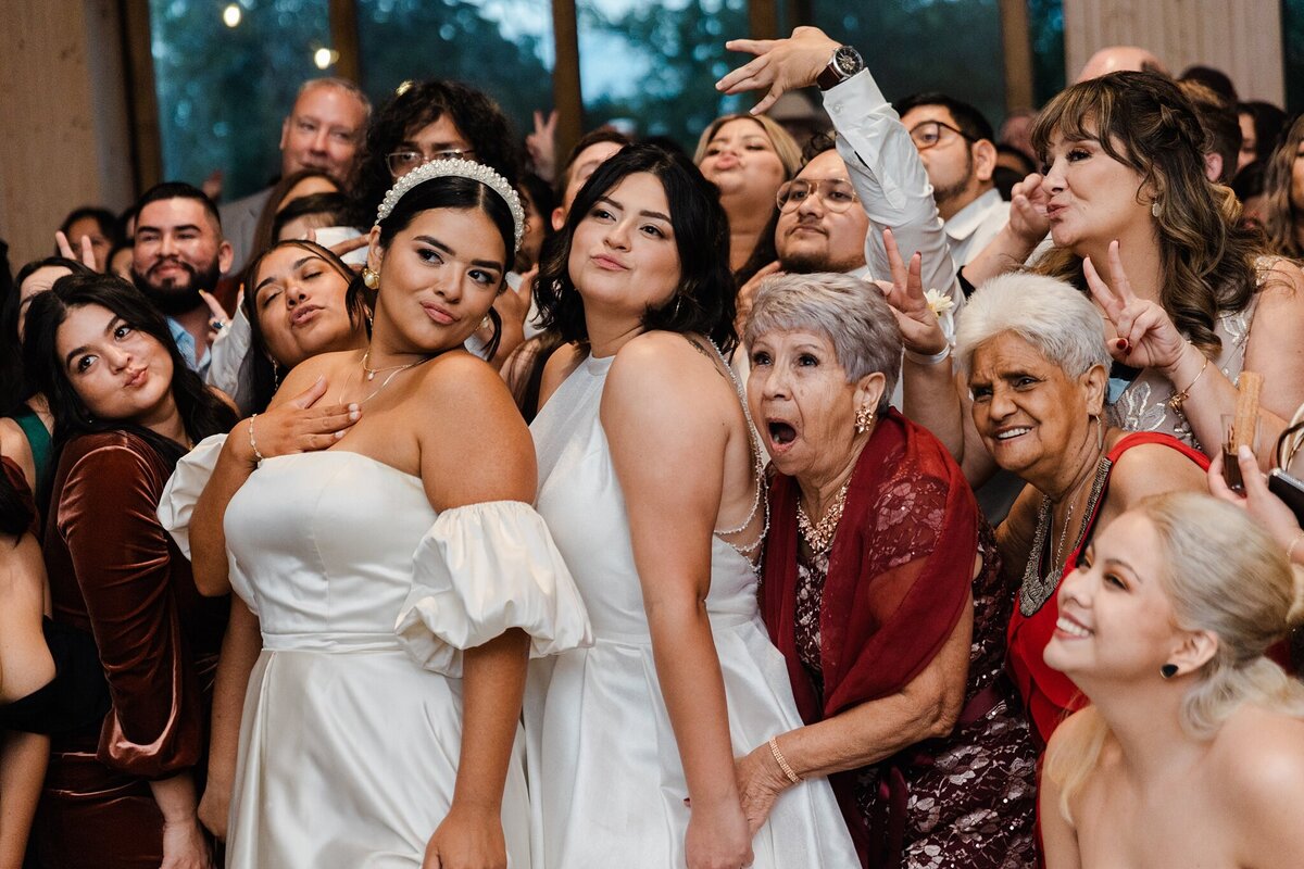 Candid shot of two brides and many of their wedding guests and family posing for another camera during their wedding reception in Fort Worth, Texas. Both brides are wearing sleeveless, elegant, white dresses while the bride on the left is also wearing a highly decorated headband.