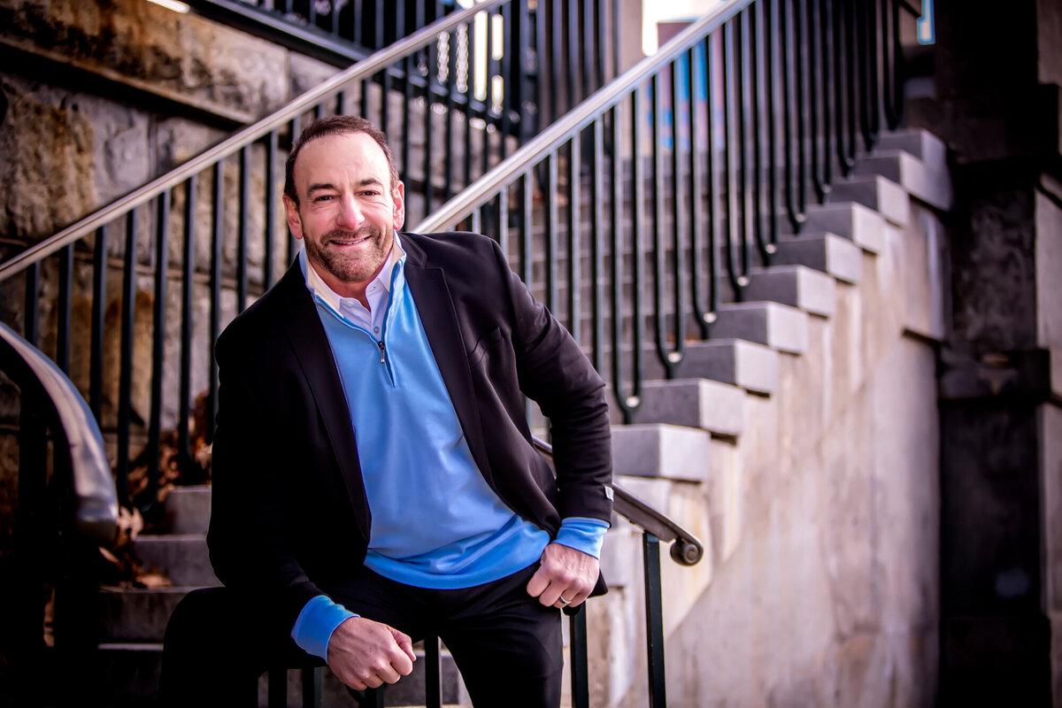A male physician standing at the bottom of an outdoor stone staircase, wearing a blue sweater & blazer, posing for a South Shore MA branding photographer
