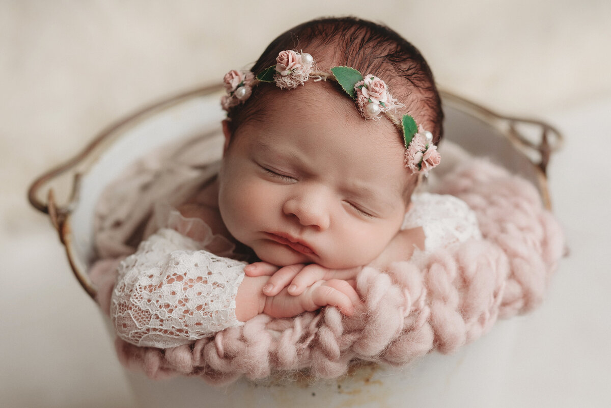 1 week old newborn girl posing in white bucket with lace outfit and flower headbands with head on chin