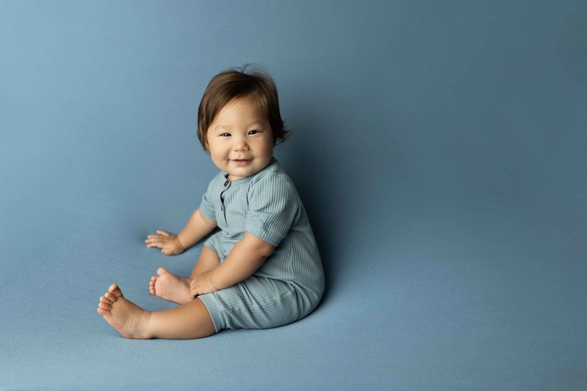 Baby in Blue onesie sitting on a plush, blue infinity backdrop. Baby facing the side but looking and smiling at the camera.