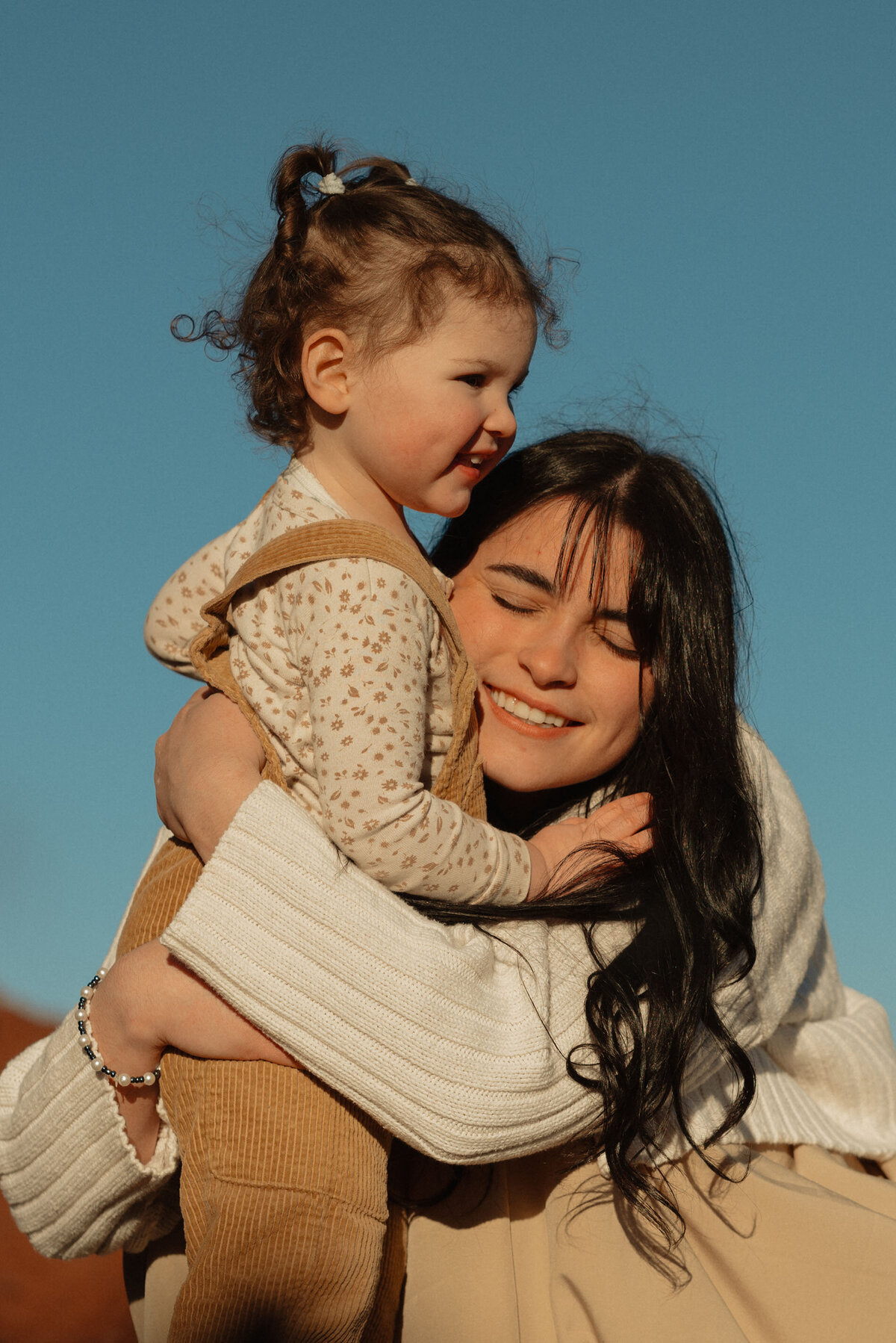 mom smiles and holds daughter while standing against the blue sky in central Oregon