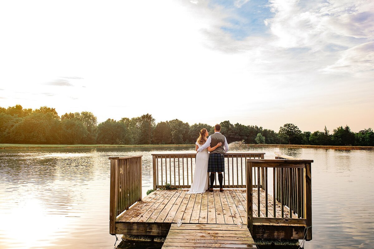 Standing on a dock in the middle of a lake, the bride and groom embrace as they watch the sunsetting into the trees at their Nashville elopement. The bride is in a fitted off the shoulder wedding dress with a short train. The groom is wearing a gray vest with a blue and green kilt with tall gray socks and brown shoes.