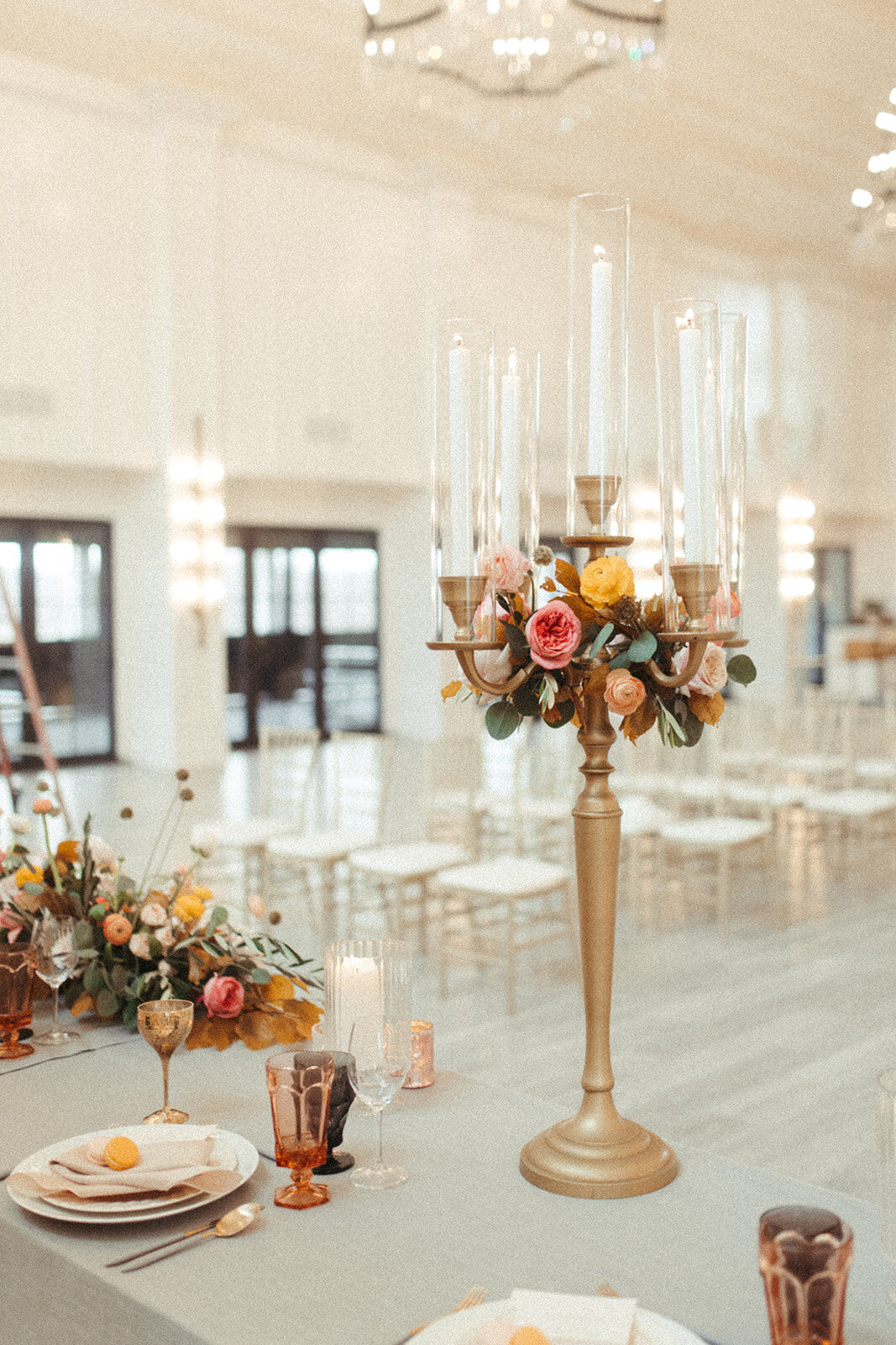 A table set with gold gray linen, candlesticks, flowers, set against a room filled with white and gold chairs.