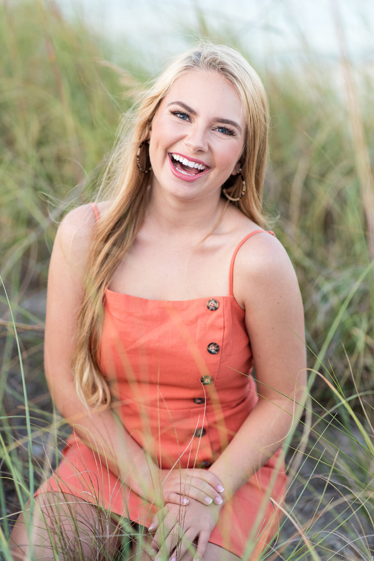 Clover Hill high school student laughs during her senior portrait session while kneeling in tall beach grass at Yorktown Beach, VA.
