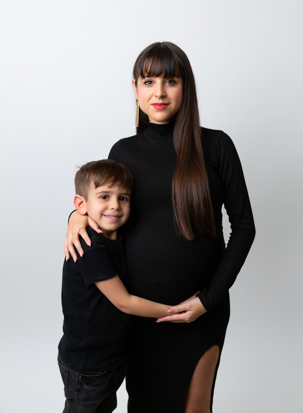 Expectant mom and older son pose for maternity photos in Brooklyn, NY photo studio. Big brother is cuddling mom and holding her bump. Mom has arms wrapped around her older son. Both are smiling at the camera.