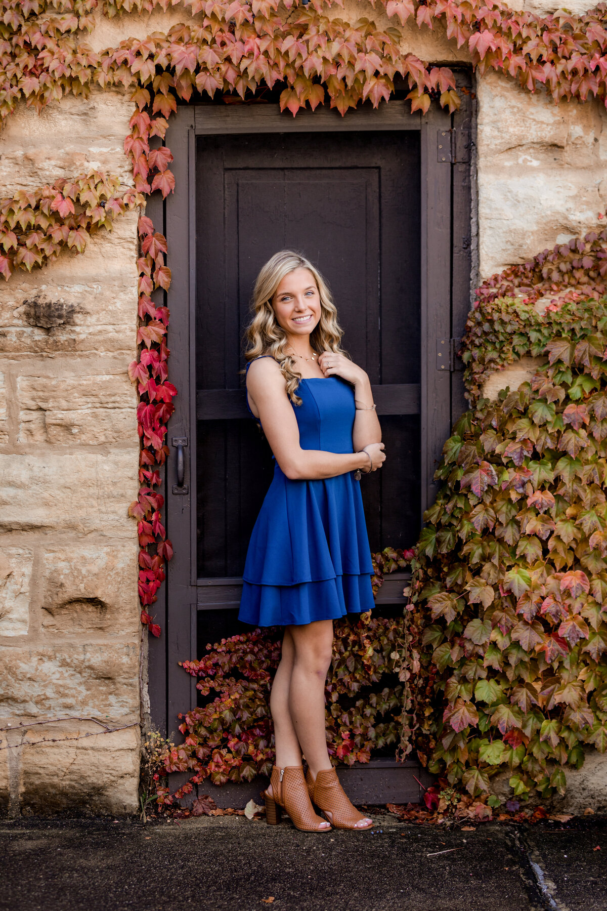 A blonde senior wears a blue dress while standing in front of a door surrounded by fall colored ivy with her ankles crossed.
