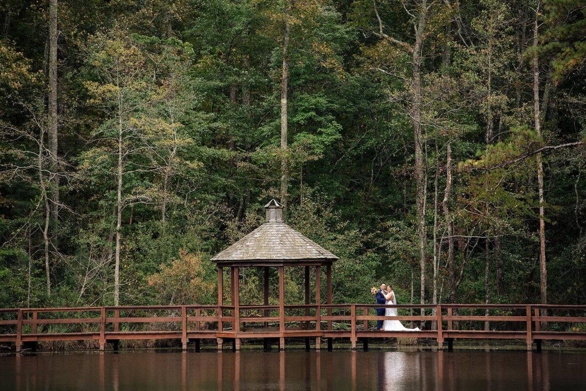 Bride and groom small in the frame standing on a bridge with their reflection in the water in Raleigh NC by Charlotte wedding photographers DeLong Photography