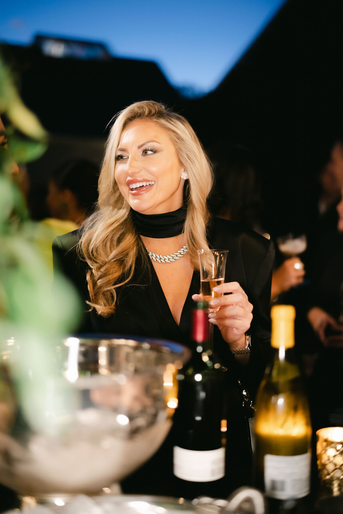 The photo was taken at a tented wedding in Virginia, beautifully captured by photographer Elisabetta Marzetti, with videographers Clayton Films also present to document the special day. In the image, a woman with long blonde hair, dressed in a black outfit and a silver necklace, is seen smiling as she holds a glass of beer. She is positioned near a table adorned with wine bottles and an ice bucket, with other guests appearing blurred in the background. The overall ambiance of the setting suggests that the event took place in the evening, creating a warm and inviting atmosphere.