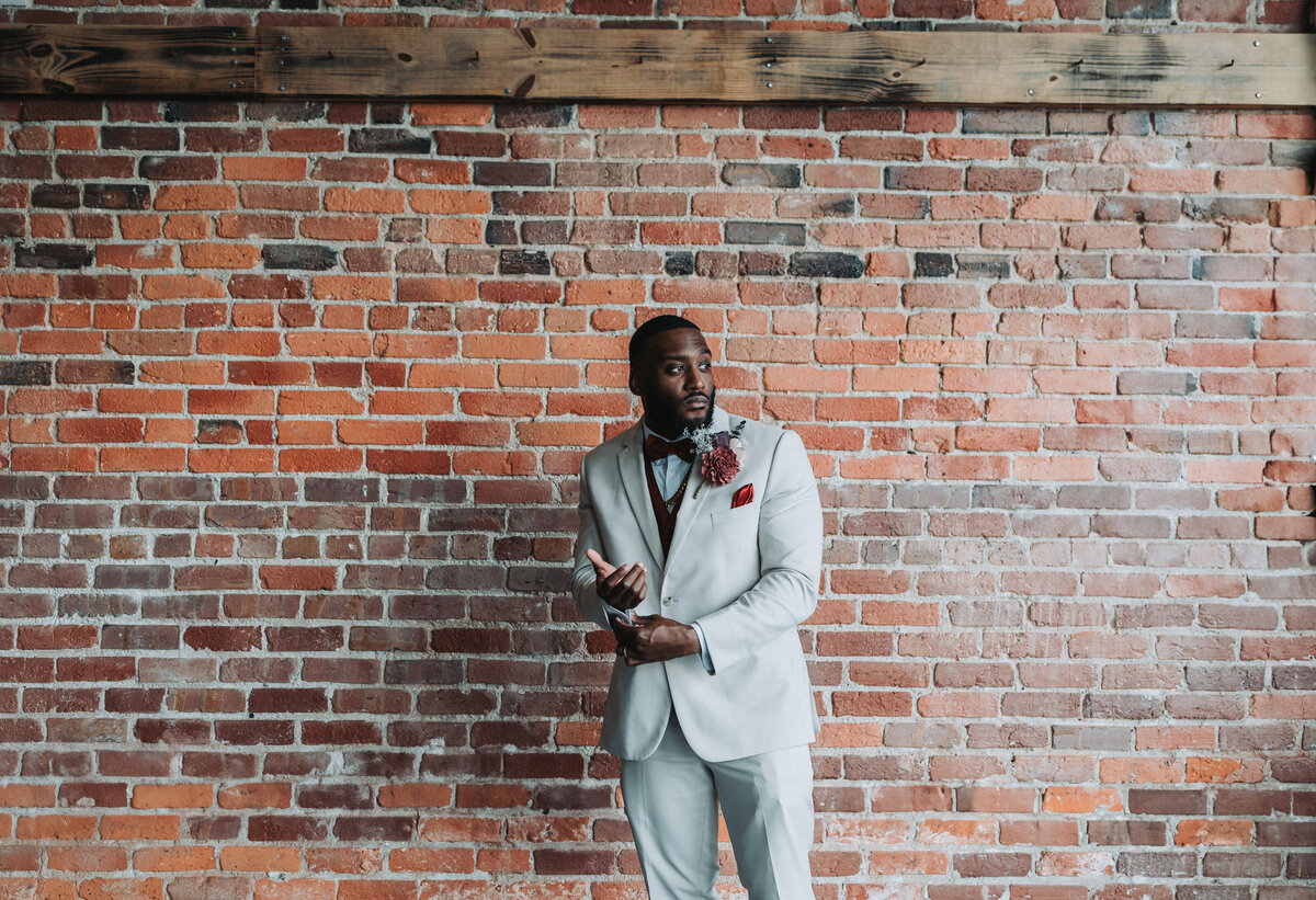 A stylish groom in a light-colored suit with a burgundy bow tie and boutonniere poses confidently against an exposed brick wall at the industrial-chic Neon House in Norfolk, Virginia, captured by Quinn Photography LLC.