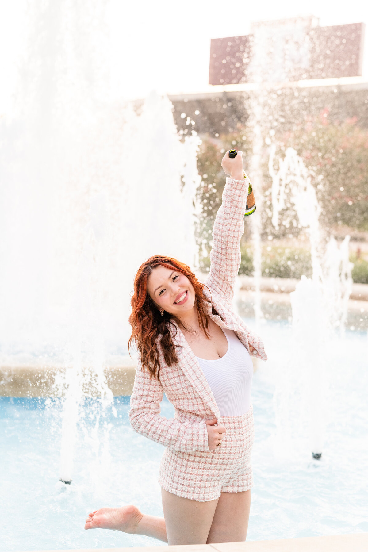 Texas A&M senior girl wearing plaid shorts and blazer while standing in pond and smiling and holding up champagne bottle in front of Kyle Field