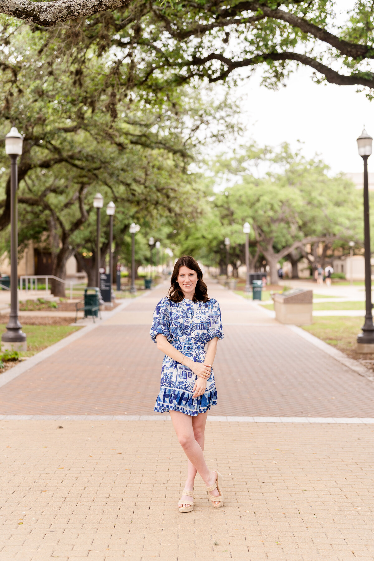 Texas A&M senior girl standing in blue and white dress while holding wrist and smiling under the trees of Military Plaza