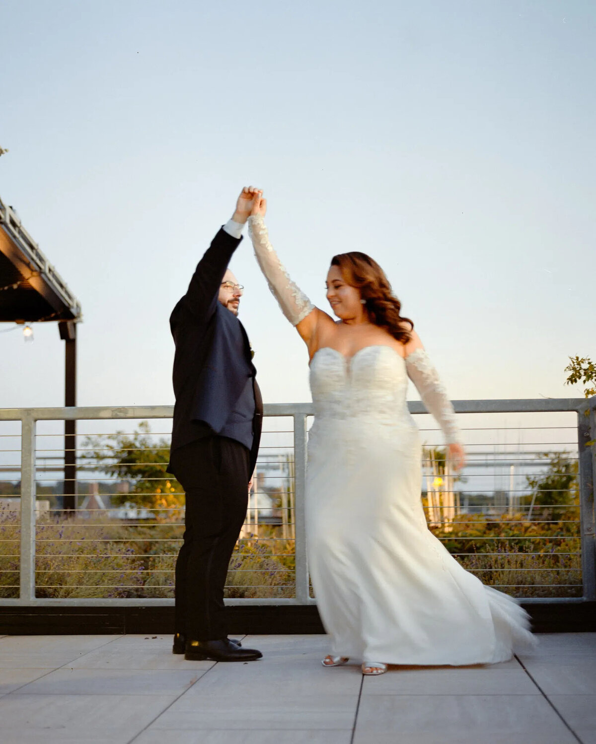 A couple dressed in formal wedding attire dance on an outdoor terrace at sunset. Captured by an NJ wedding photographer, the person in a suit lifts their partner’s arm, twirling together in a moment full of joy and romance.
