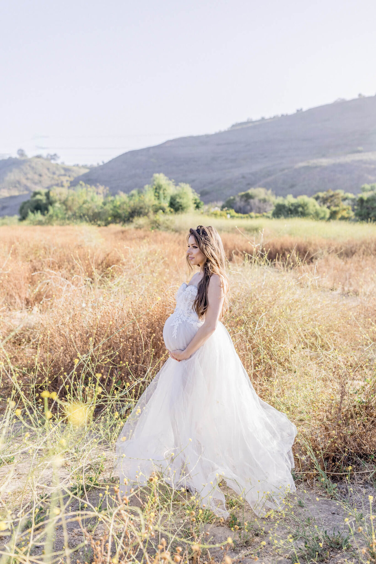 A pregnant woman in a flowing white dress stands in a sunlit field, gently holding her belly. The background features dry grass and distant hills under a clear blue sky.