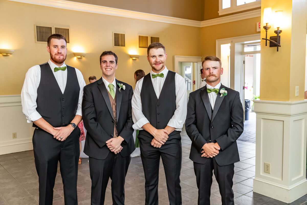 Four men dressed in formal suits with green accents stand side by side indoors, smiling for a photo at a wedding.