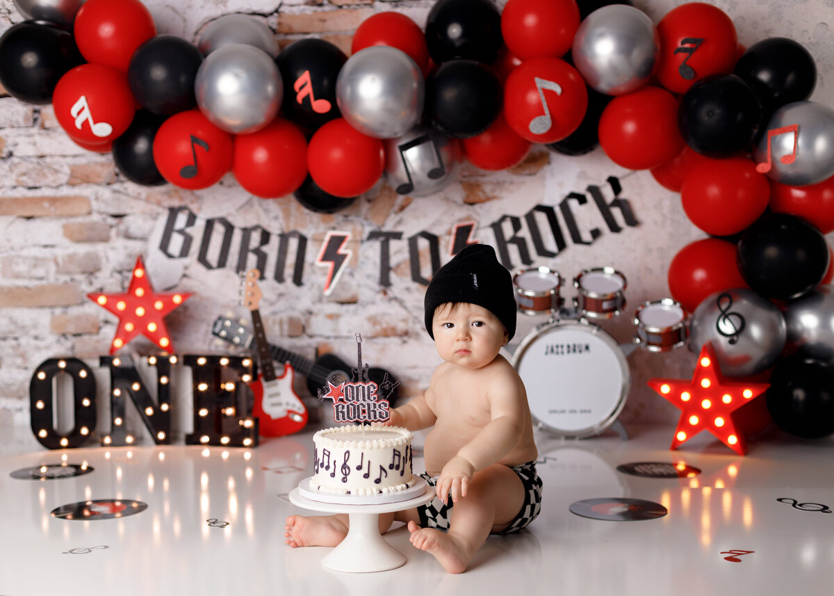Rock and roll cake smash in West Palm Beach and Delray Beach portrait studio. Baby is sitting behind a white musical note cake. In the background is a balloon garland with red, black and silver balloons, a mini drum set and guitar with record albums scattered on the floor behind a brick backdrop.