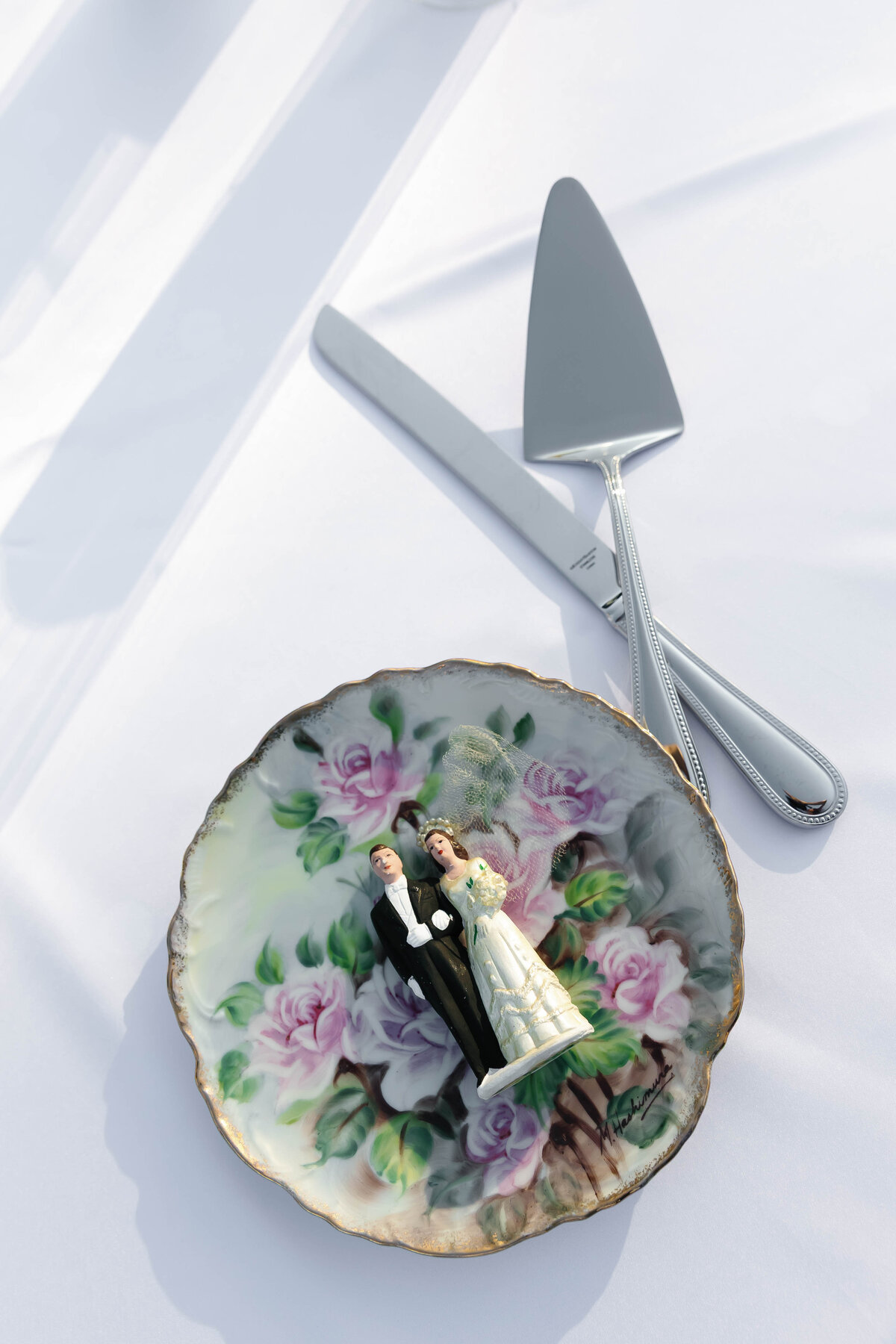 Vintage floral china plate holding a traditional bride and groom cake topper beside silver cake-cutting utensils at an outdoor wedding reception.