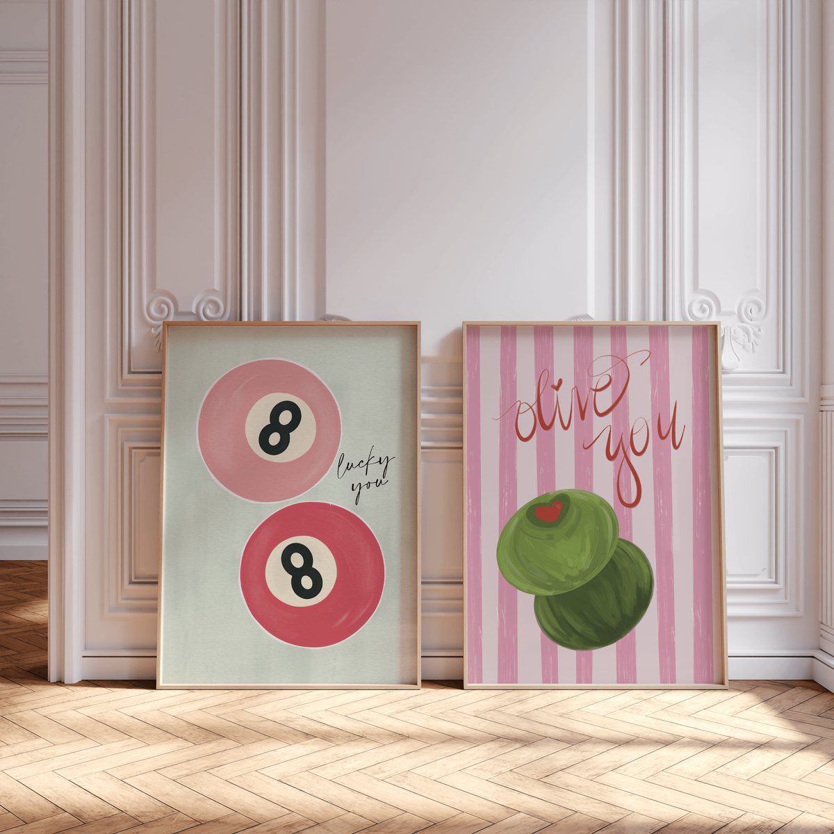 Two framed posters on a wooden floor: one by Sweet Pea Ink Designs with two pink billiard balls and text, the other with two green olives and "olive you," set against a white paneled wall. Perfect for playful brand design lovers.