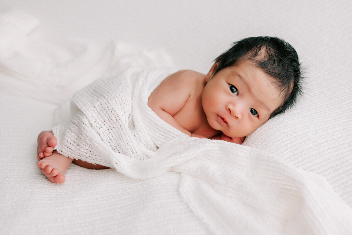 portrait of boy looking at camera while laying on white fabric for his newborn photography session