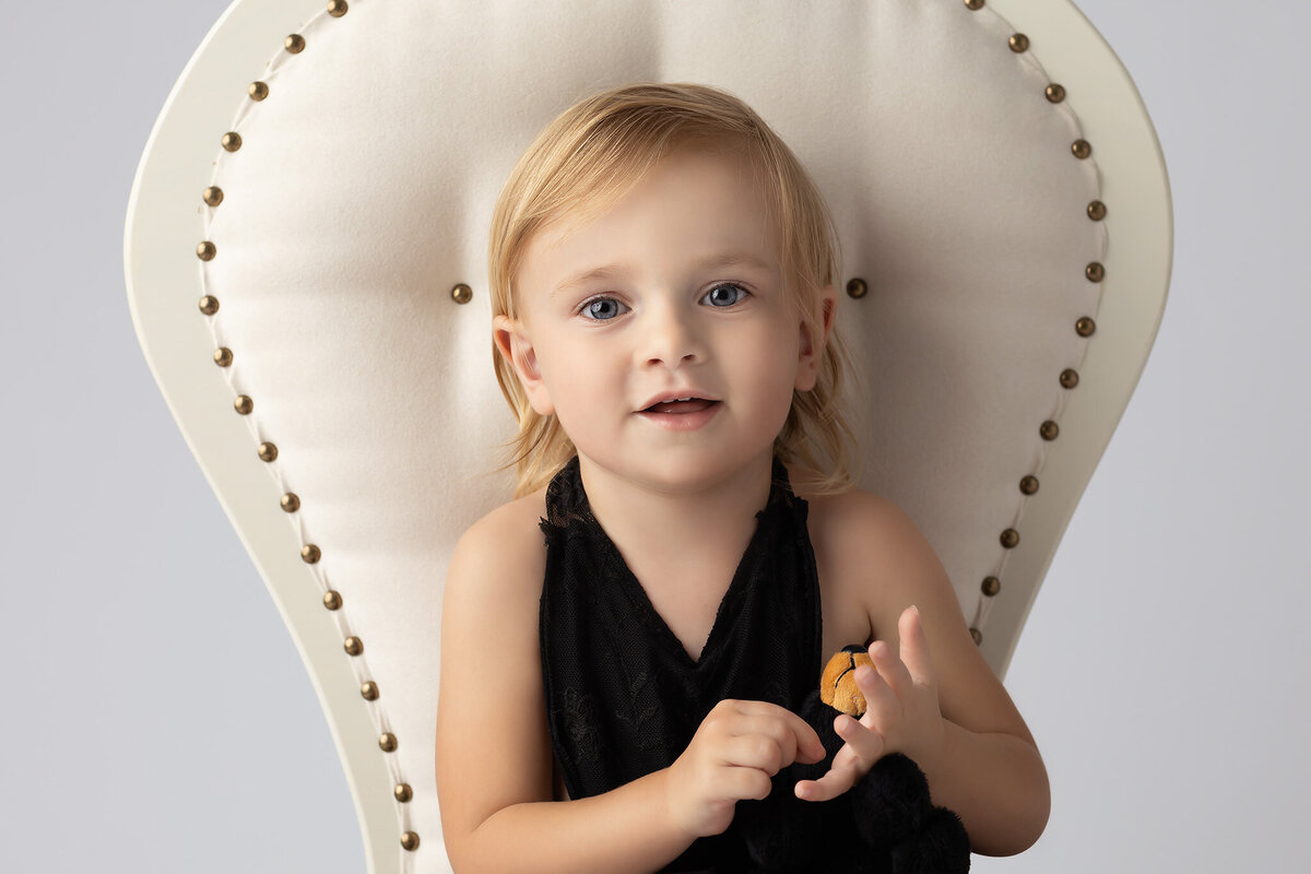 Close-up of a toddler smiling while seated in a vintage chair, holding a stuffed animal, captured by a Dallas family portrait photographer.