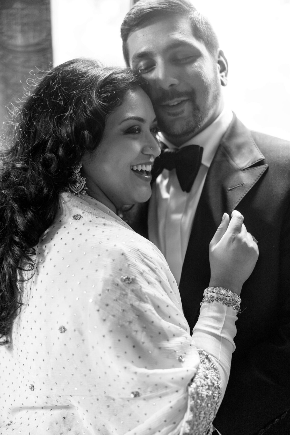 A black and white photo of a smiling couple embracing. The woman is wearing a saree with intricate beadwork, while the man is in a suit with a bow tie. They appear joyful and close, with the woman on the left and the man on the right.