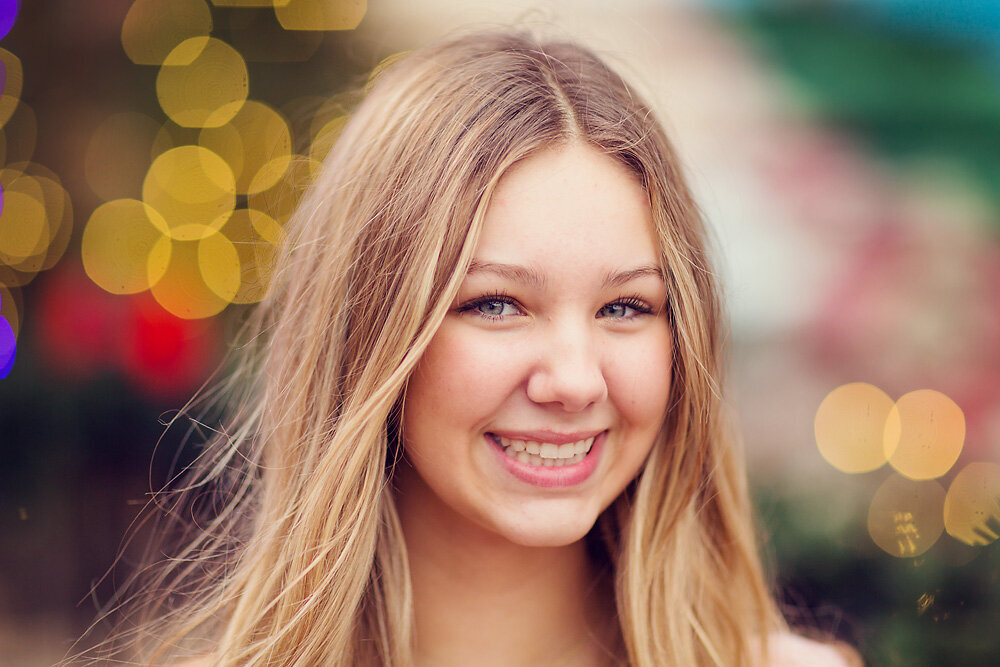 Close headshot of teenage girl smiling with Christmas lights behind her.