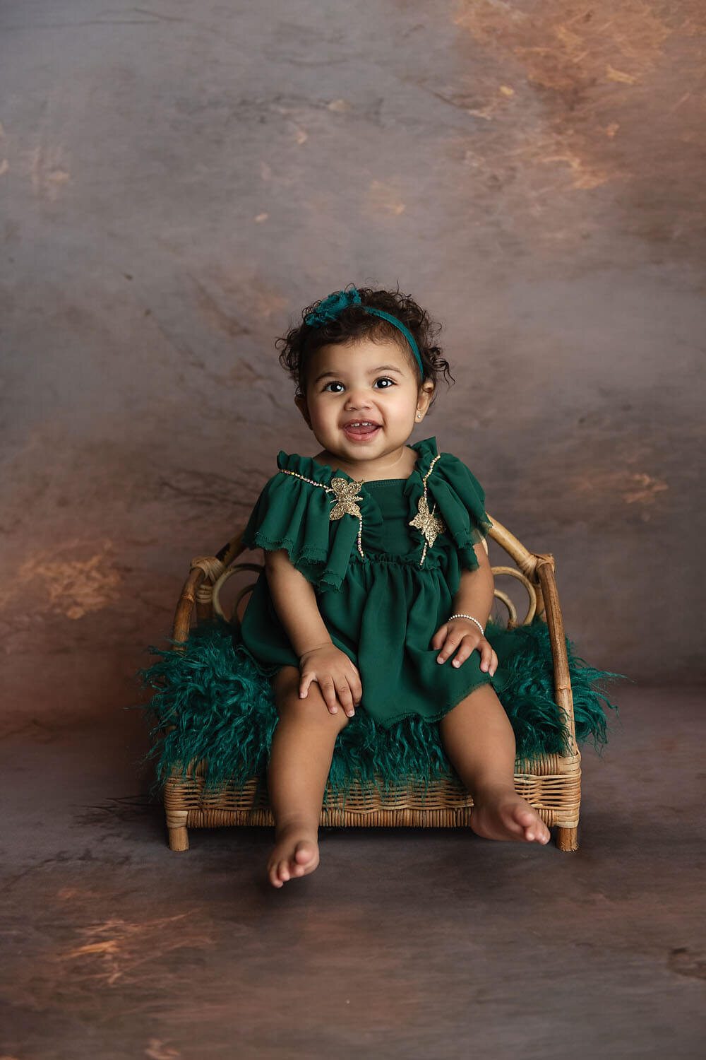 A happy toddler girl sits on a wicker chair in a green dress and headband