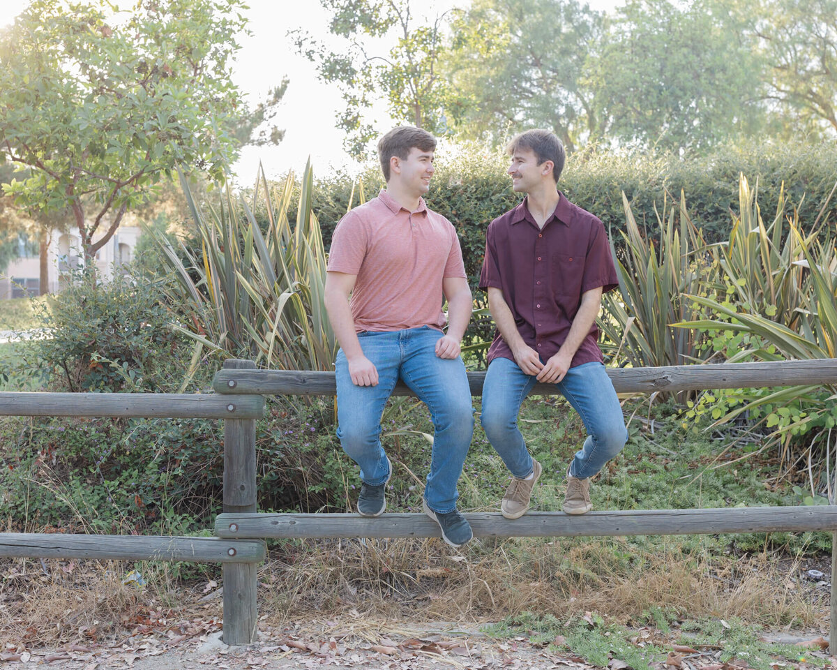 Two brothers with dirty blonde hair wearing shades of red and blue jeans, looking at each other and sitting on the top rung of a wooden park bench, with trees and greenery behind them.