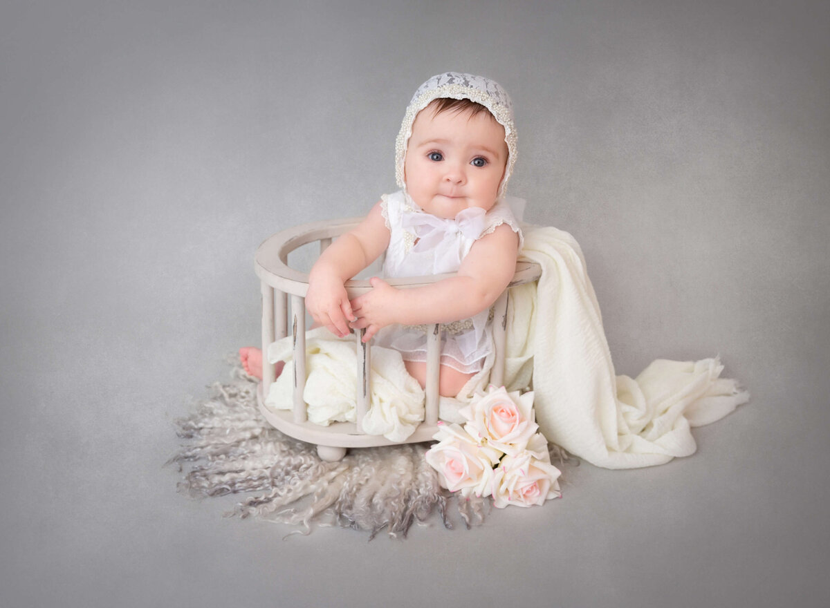 Baby girl wearing a lace outfit and matching bonnet, gazing at the camera during her 6-month milestone photoshoot by a professional NYC baby photographer