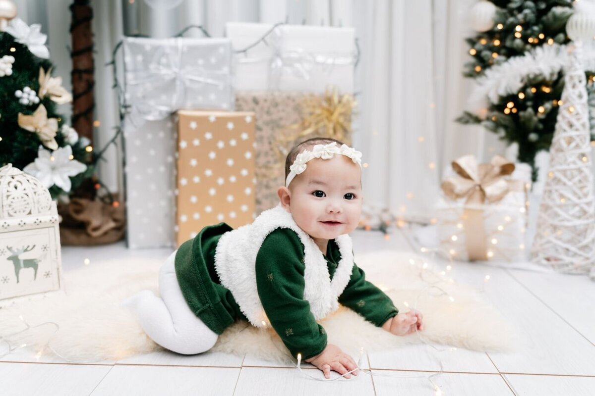 Baby girl in a green top and white headband crawling on a fluffy rug with Christmas lights and wrapped gifts.