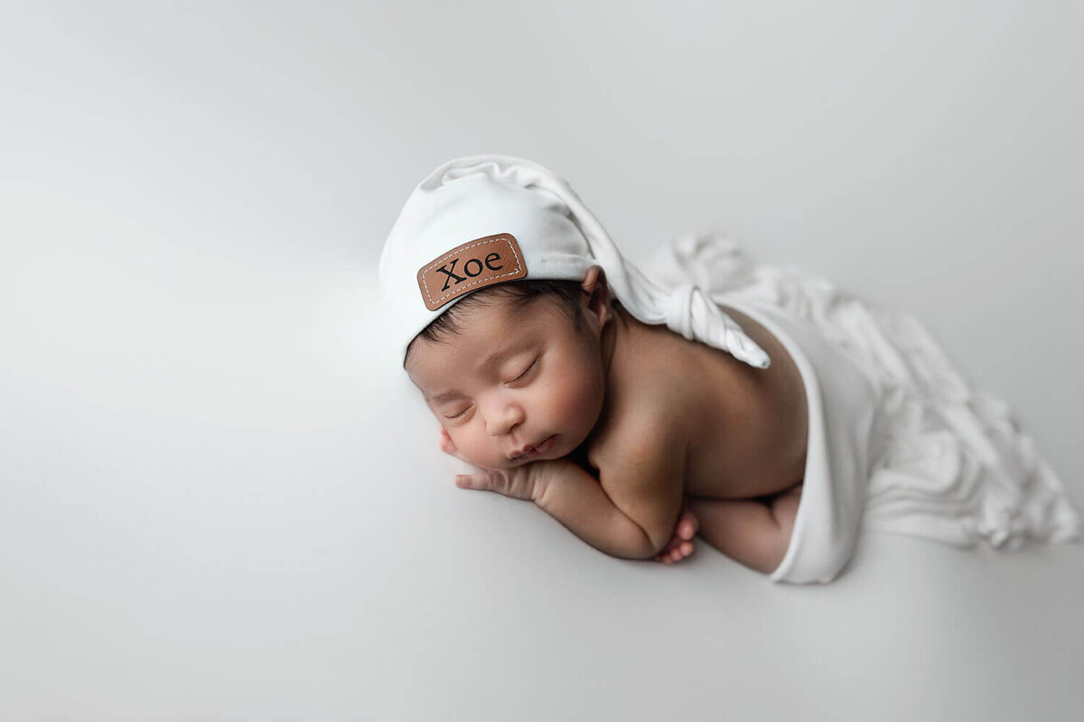 A newborn baby in a white night cap sleeps on a white pad
