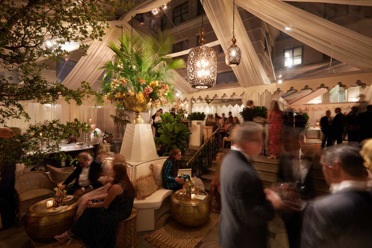 This stunning photo captures a bustling evening event at The Willard Intercontinental in Washington D.C., one of the top wedding venues and hotels in the city. Captured by photographer Shawn Connell, the indoor space is beautifully decorated with draped fabric and ornate hanging lights, creating an elegant setting. Elegantly dressed people can be seen socializing amidst lush greenery, gold tables, and ambient lighting, all contributing to a warm and inviting atmosphere.