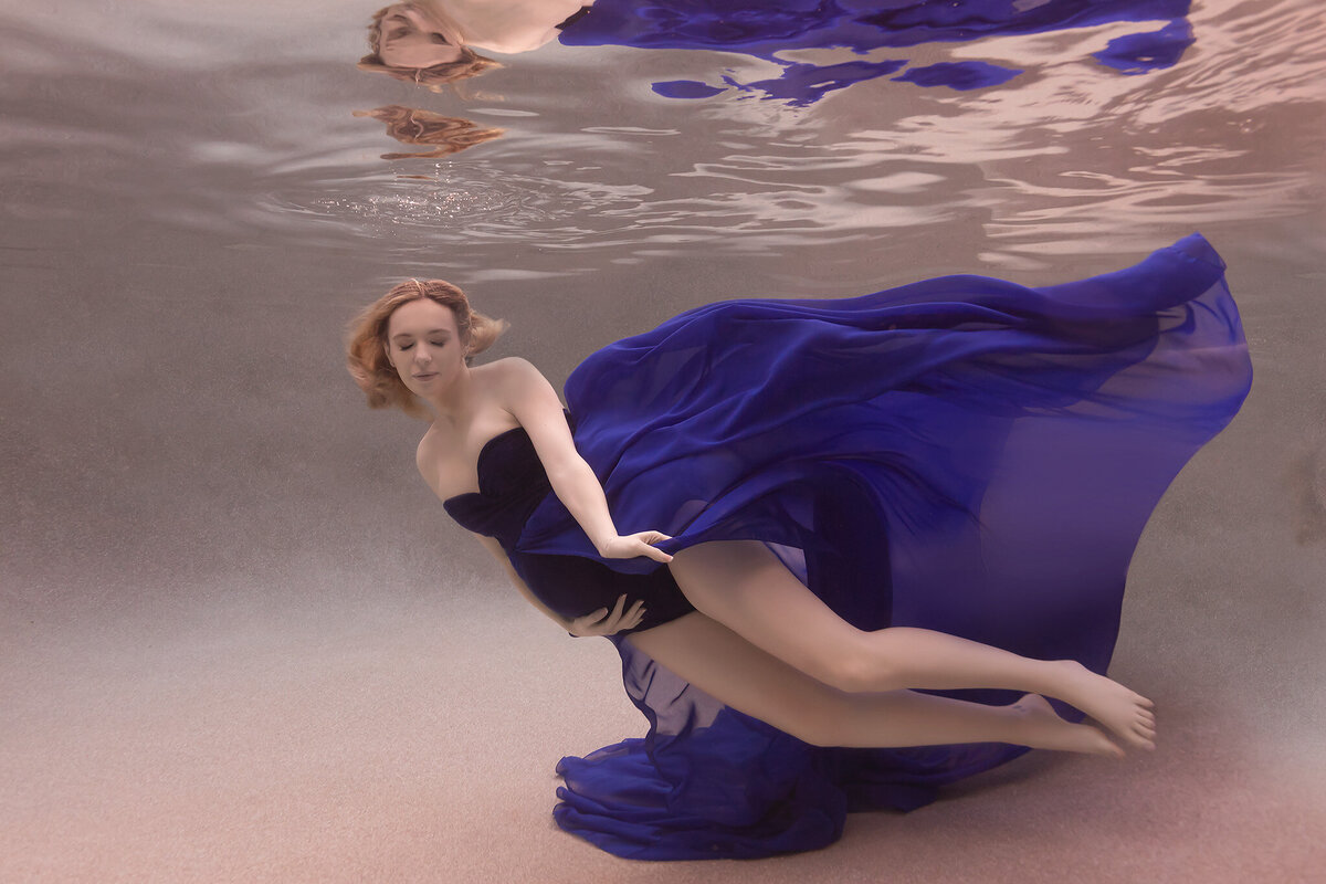 A graceful underwater maternity photo taken by a Dallas underwater maternity photographer. The woman is elegantly posed, her deep blue gown flowing around her as if suspended in motion. Her eyes are gently closed, adding to the sense of peacefulness and ethereal beauty. This underwater maternity photoshoot highlights the weightlessness and tranquility of the experience, enhanced by the soft light filtering through the water.
