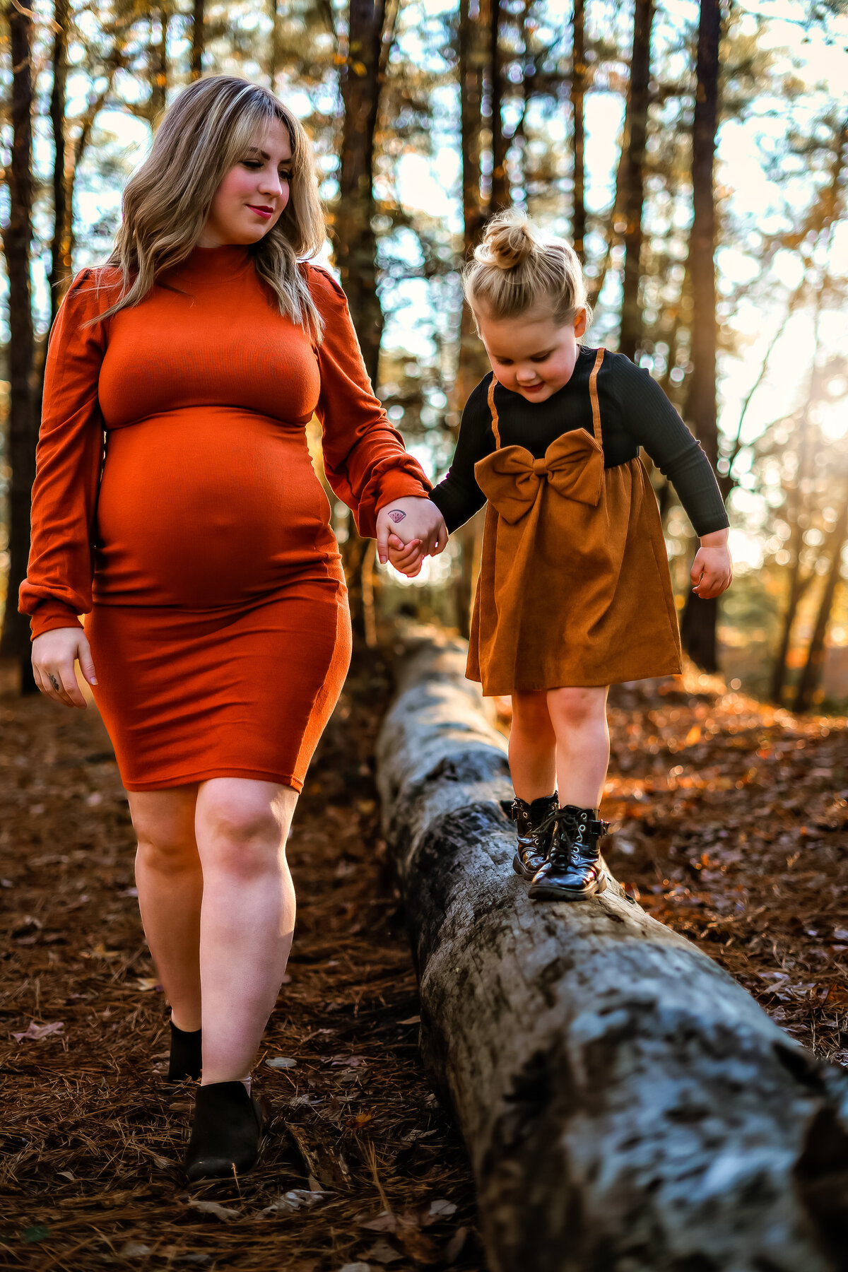 Expectant mother holding daughters hand while she walks on a log wearing fall colors