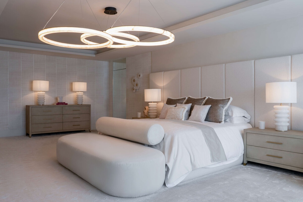 A spacious primary bedroom with a focus on light, neutral tones. Above the bed is a contemporary chandelier composed of three circles, emitting a soft warm light through the room. Lamps on the bedside tables and dresser emit a similar light.