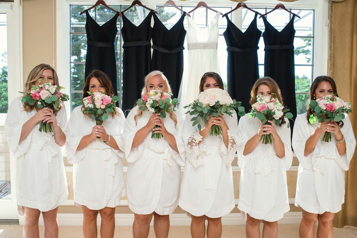 Bride and bridesmaids in robes holding bouquets in front of their faces with dresses hanging in window at Stonebridge Country Club