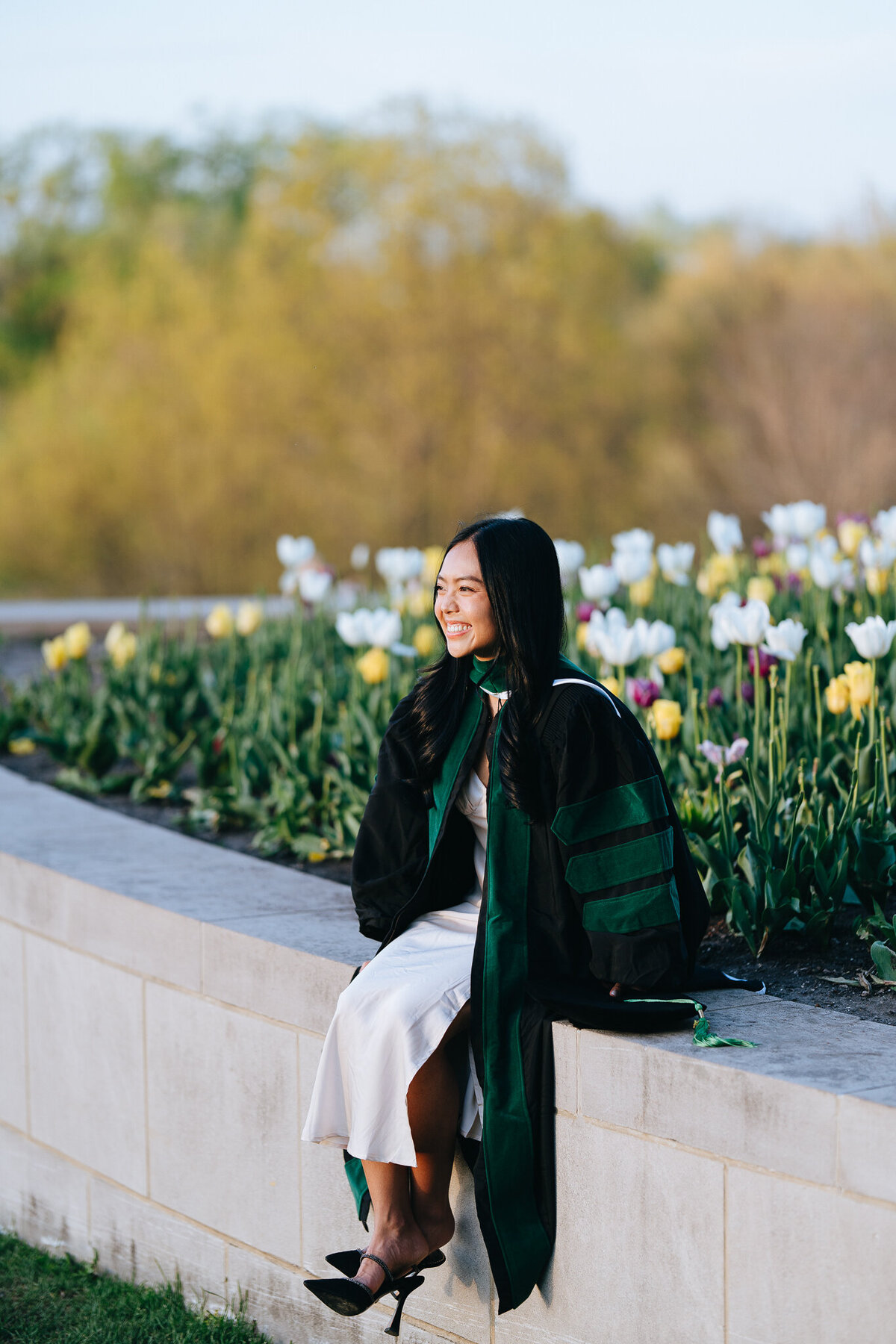 slu med grad wearing regalia sitting by bed of flowers in forest park