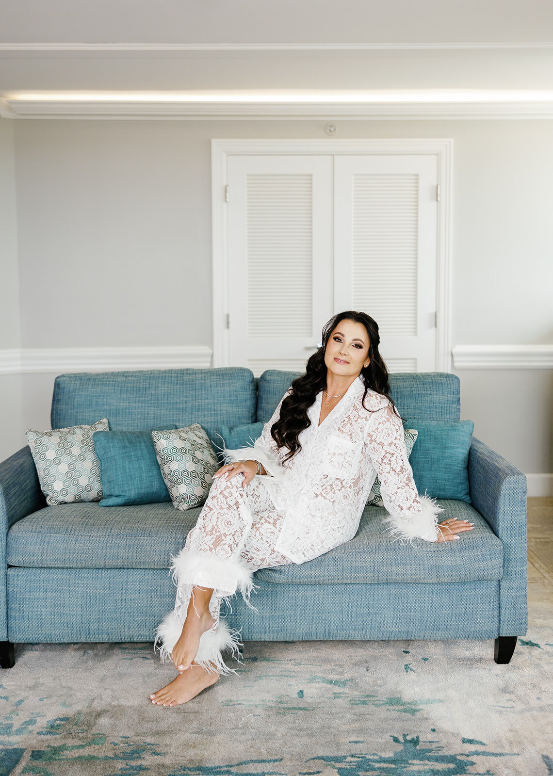 The bride sitting gracefully on a blue couch in her white lace pajamas, enjoying the quiet moments before the ceremony at Ashley and Cameron's micro wedding at Ocean Reef in Key Largo, captured by Claudia Amalia Photography.